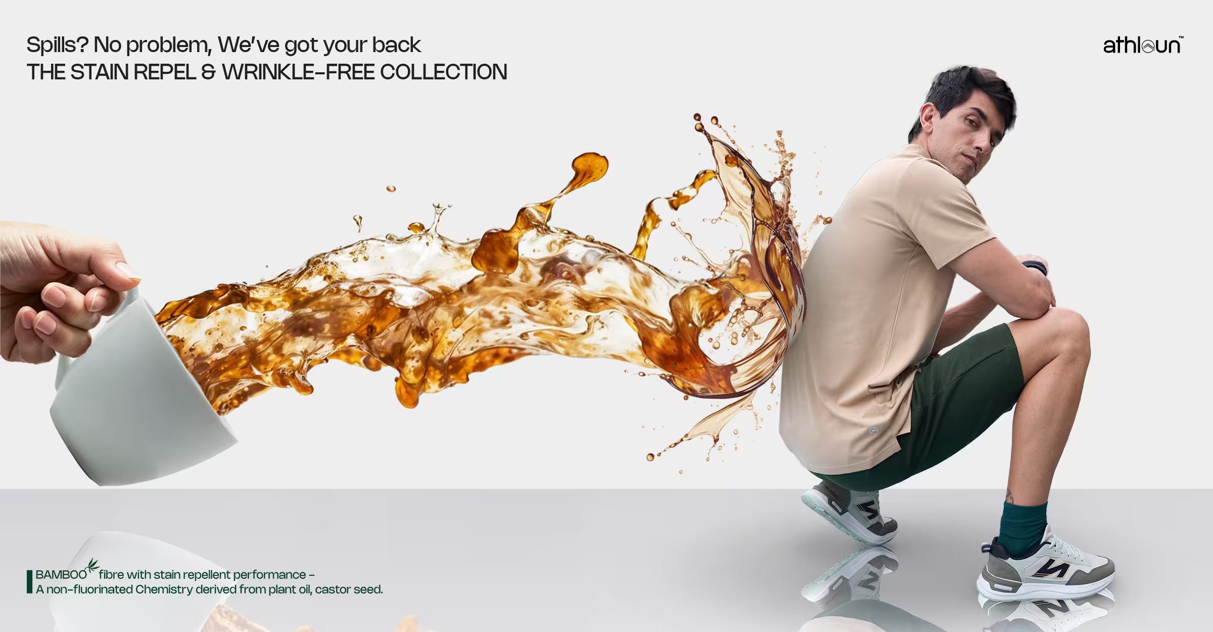 A promotional ad-style image showing a man crouching on a white studio floor while wearing a beige T-shirt and dark green shorts. A hand at left holds a white mug, and a large splash of amber-brown liquid arcs across the center, suggesting spill resistance.