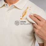 Close-up of a cream-colored polo shirt with a brown stain on the chest, being wiped with a white cloth. The shirt features a black 'STAIN-REPELLENT' logo and icon on the chest.