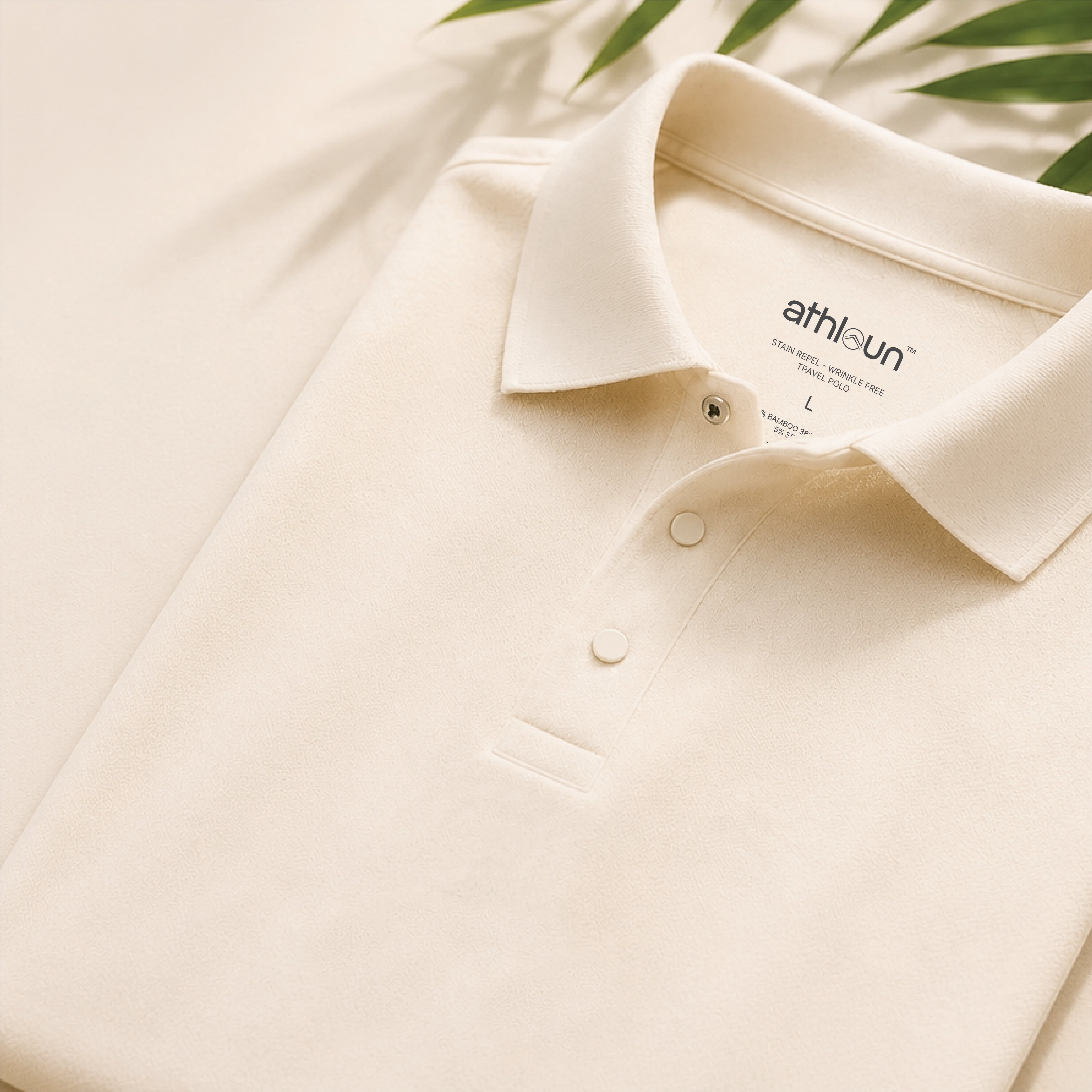 An off-white polo shirt folded neatly, featuring a classic collar, two-button placket, and a visible 'athlaun' brand label inside the collar. The shirt is styled with soft green palm leaves on a matching light background.
