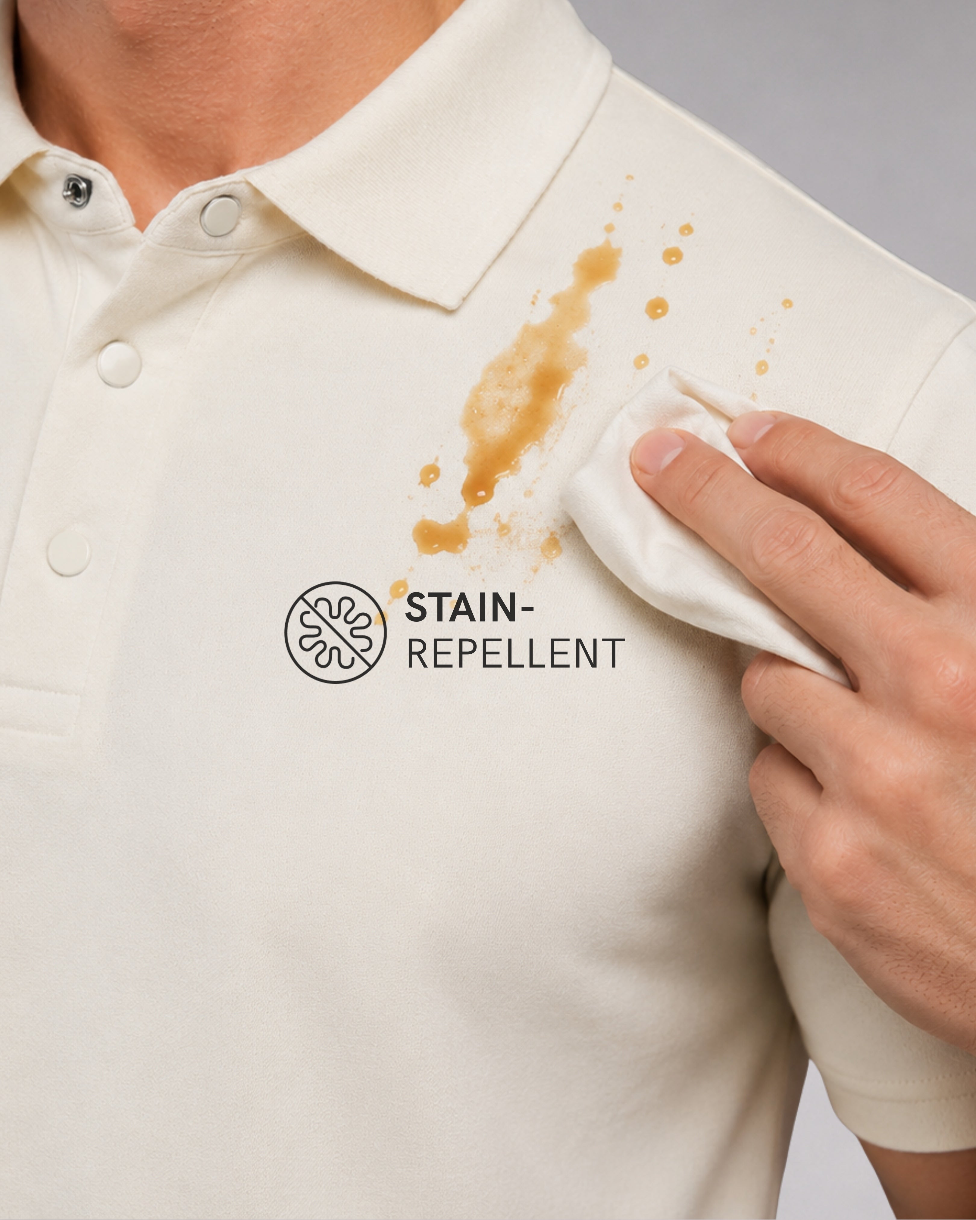 Close-up of a person wearing a cream-colored polo shirt with a brown liquid stain on the chest and a hand rubbing a stain-repellant area. The text 'STAIN-REPELLENT' is printed below the collar.